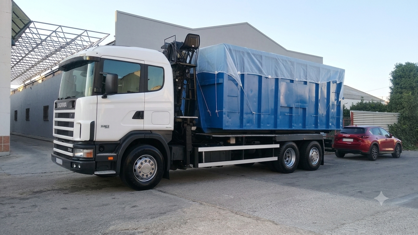 Camión blanco con caja azul cubierta, estacionado en patio industrial junto a un coche rojo.